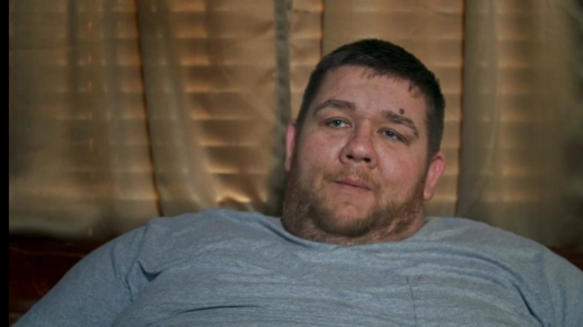Paul MacNeill on My 600-lb Life wears grey t-shirt while sitting in front of beige curtains..