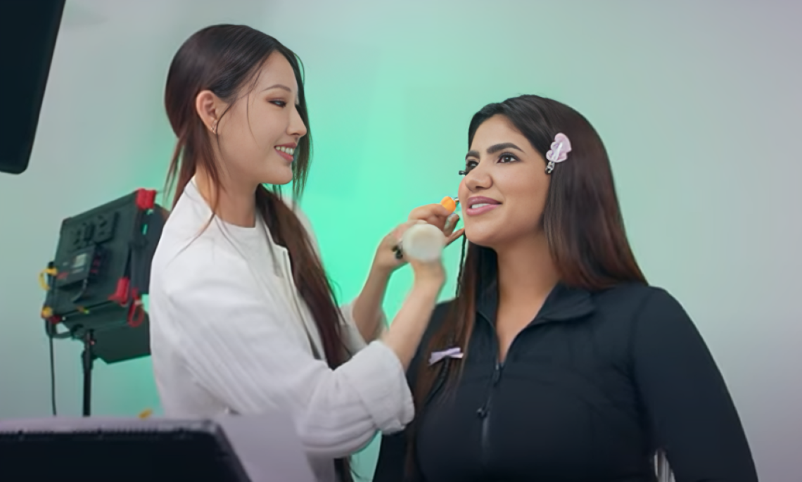 Noor Naim smiles to right of make-up artist on Super Rich in Korea, while getting make-up done.