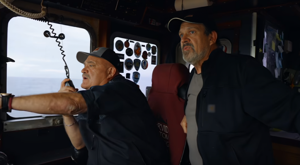 Captain Colburn speaks into mic and reaches one arm forward while sat at front of F/V Wizard to left of fellow Deadliest Catch star, as they look out of window.