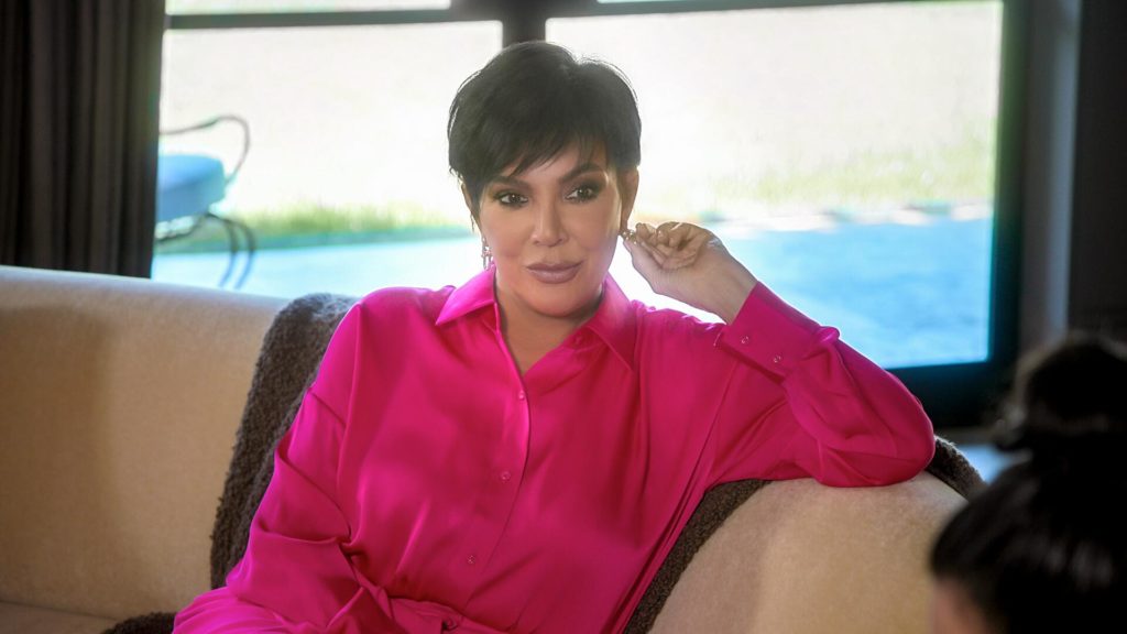 Kris Jenner wears pink shirt and touches right ear while leaning arm on sofa on The Kardashians.