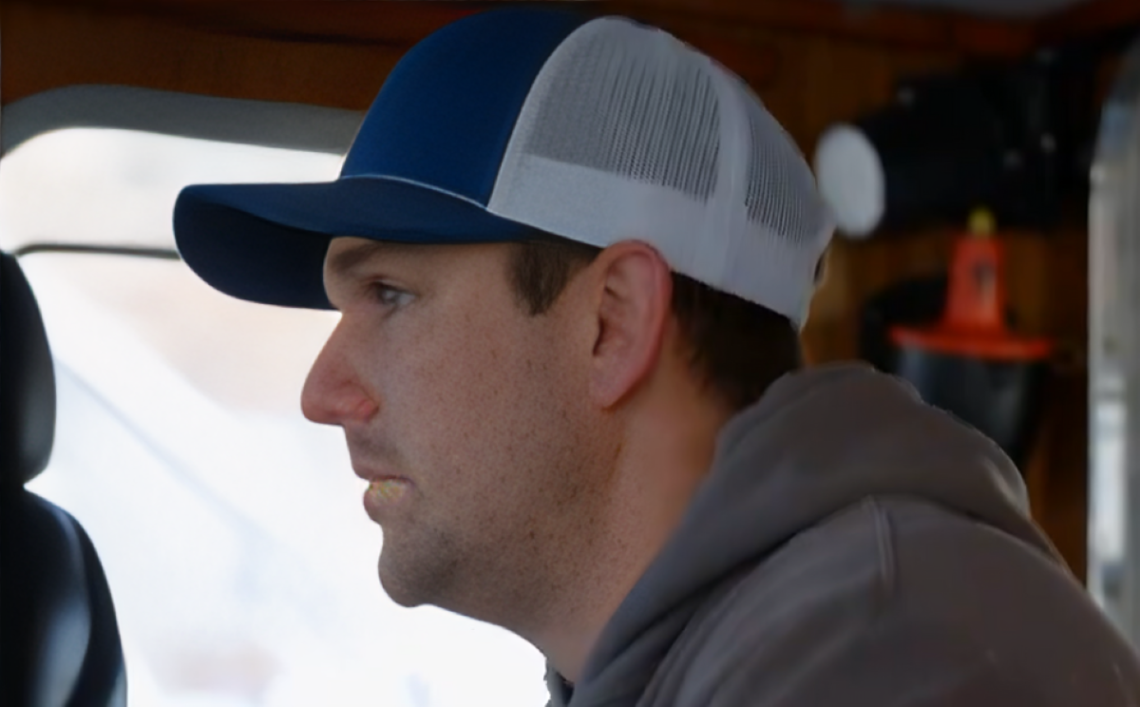 Clark faces left while wearing hat on Northwestern boat on Deadliest Catch, next to window.