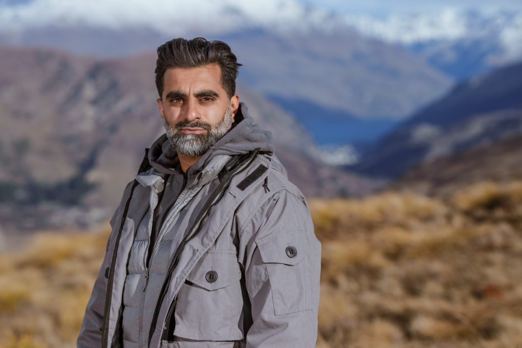 Tez Ilyas mira a la cámara con una camiseta gris y las montañas al fondo.