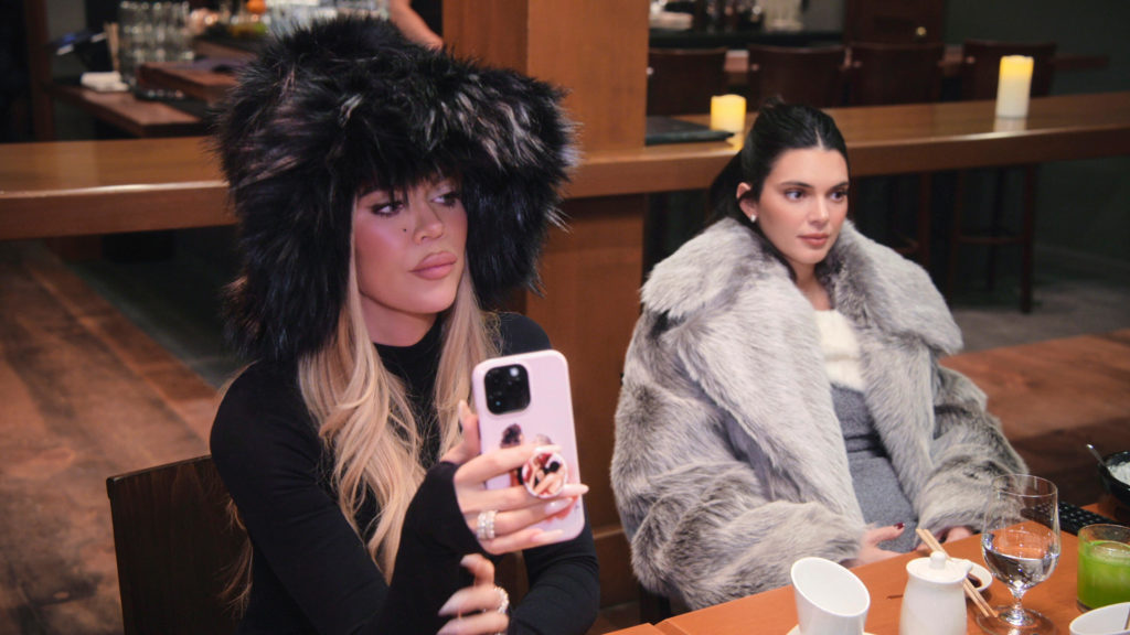 Khloé Kardashian and Kendall Jenner sit at table filming The Kardashians