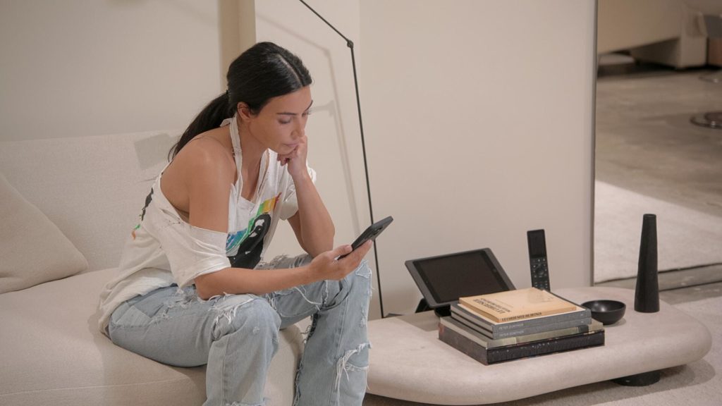 Kim Kardashian looks down at her phone sitting on couch on The Kardashians