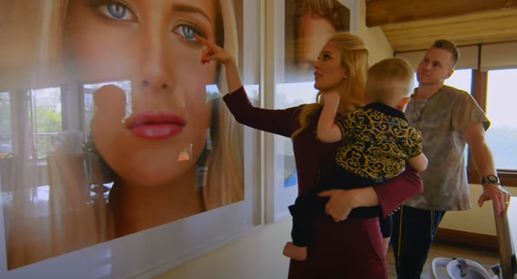 Heidi Montag points at huge portrait of herself while holding son in arms, Spencer Pratt stands behind to right.